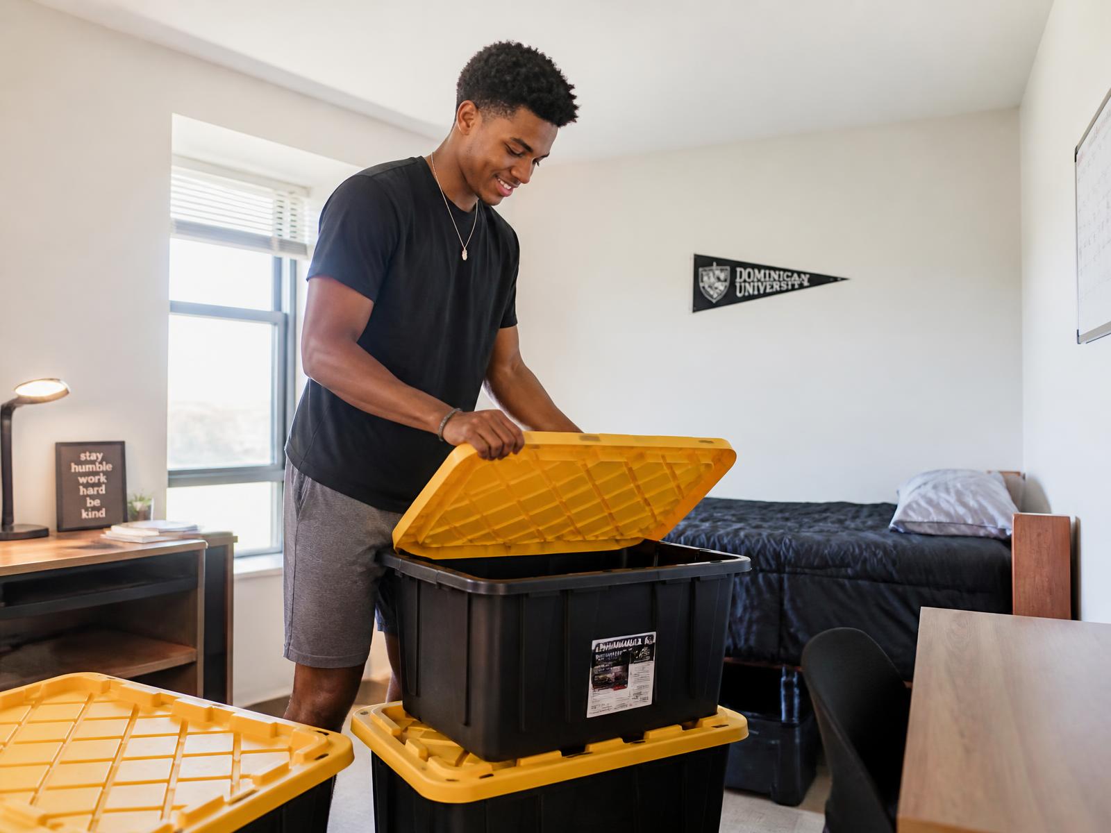 College student unpacking a stack of moving totes with yellow lids in a minimalist dorm room